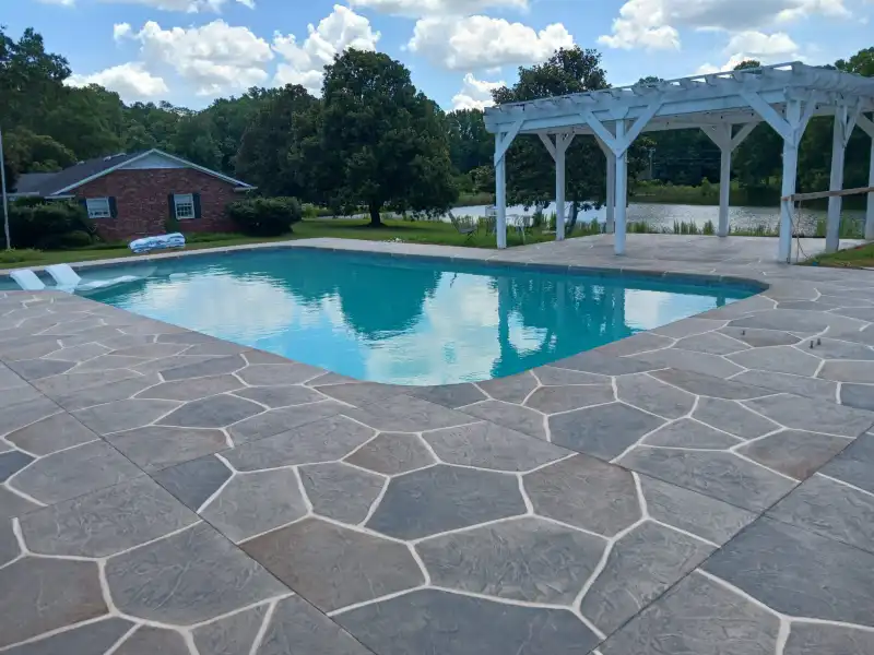 A rectangular pool surrounded by a grey and tan flagstone-pattern overlay deck, with a white pergola and a lake behind.