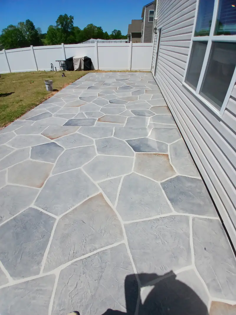 A grey flagstone-pattern overlay surface running along the side of a house between the wall and a lawn.