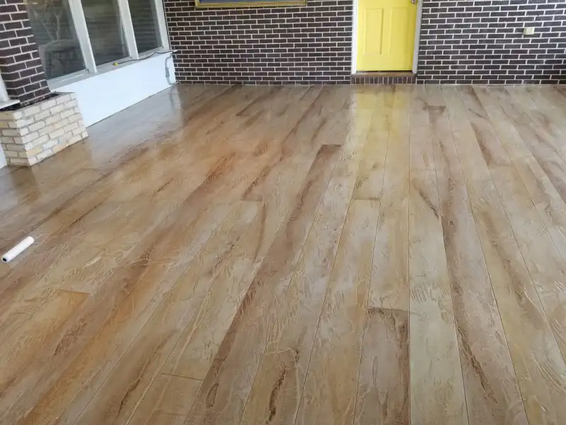 A covered porch floor finished with a wood-plank patterned overlay stained in warm brown tones, with a yellow door beyond.