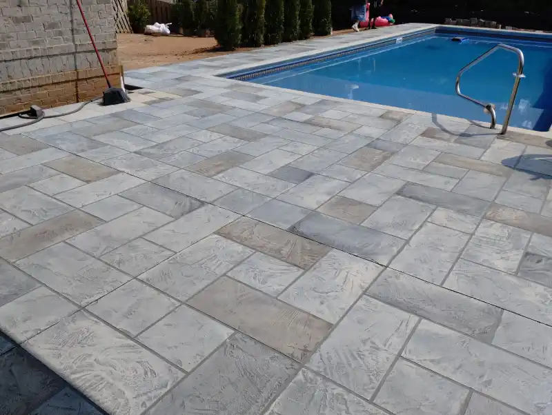 A pool deck finished with a grey rectangular tile-pattern overlay, running to the edge of an in-ground pool with a handrail.