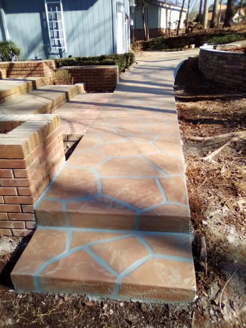 A walkway and steps finished with a terracotta-toned flagstone-pattern overlay, running between brick planters toward a house.