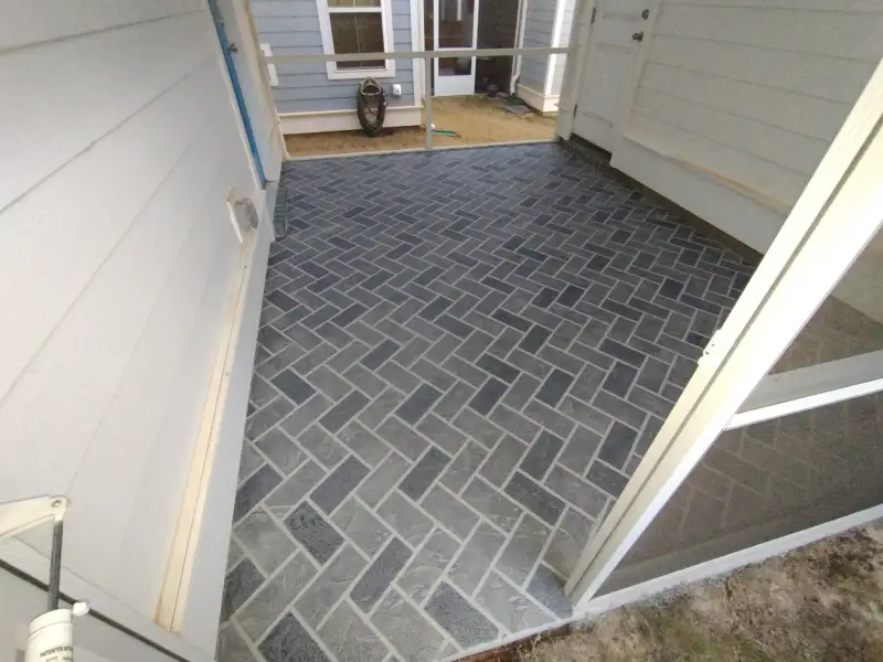 A grey mottled-texture overlay on a porch slab and adjoining walkway at a blue-clad house with a brick border.