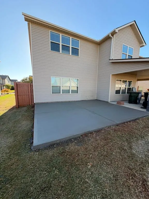 A finished broom-finished concrete patio behind a two-story house with tan siding, reaching over to the covered porch at the right, with a wood fence at the left