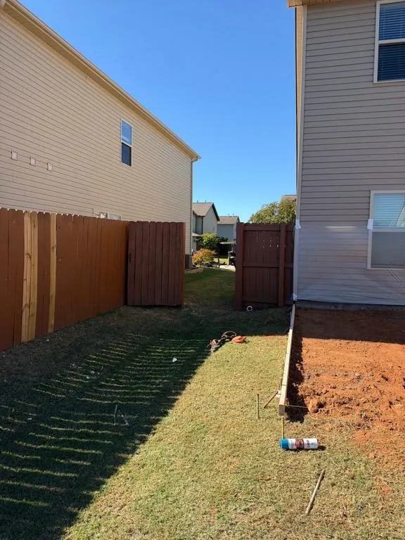 Red clay dug out next to a house with a wood form board staked along the edge, a wood privacy fence and lawn alongside
