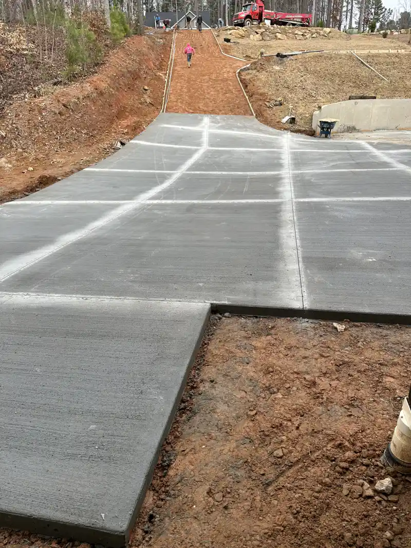 Freshly poured concrete driveway panels running up a graded cut, with a dump truck and workers at the top of the slope.