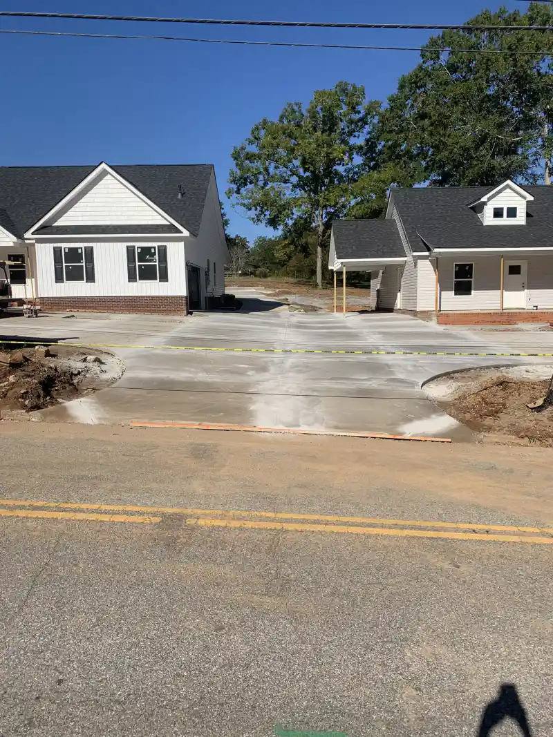 Concrete driveway aprons at two newly built houses, photographed from the opposite side of a two-lane road with caution tape across the fresh concrete.
