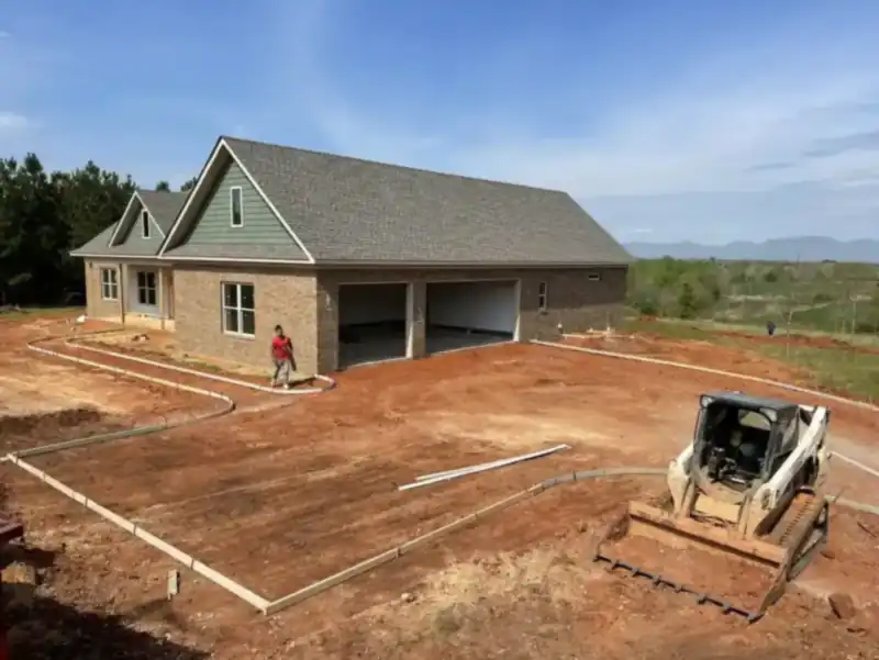 A house under construction with its garage slab graded and formed, and a skid-steer loader staged in the yard.