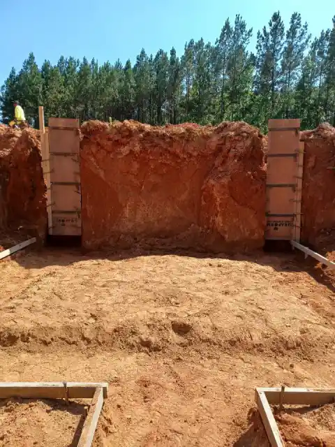 Concrete wall forms standing against an excavated red clay bank, with footing forms laid out on the pad below.