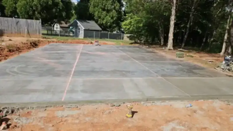 A large outdoor concrete pad freshly poured in a fenced back yard, with layout lines marked on the surface