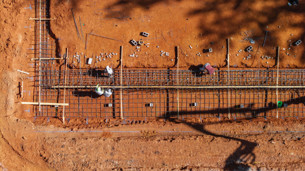 Overhead drone view of steel rebar mats and timber footing forms laid out on red clay, with workers tying steel